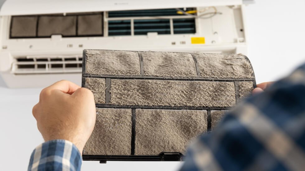 Person holding a dirty air filter in front of an open air conditioning unit.
