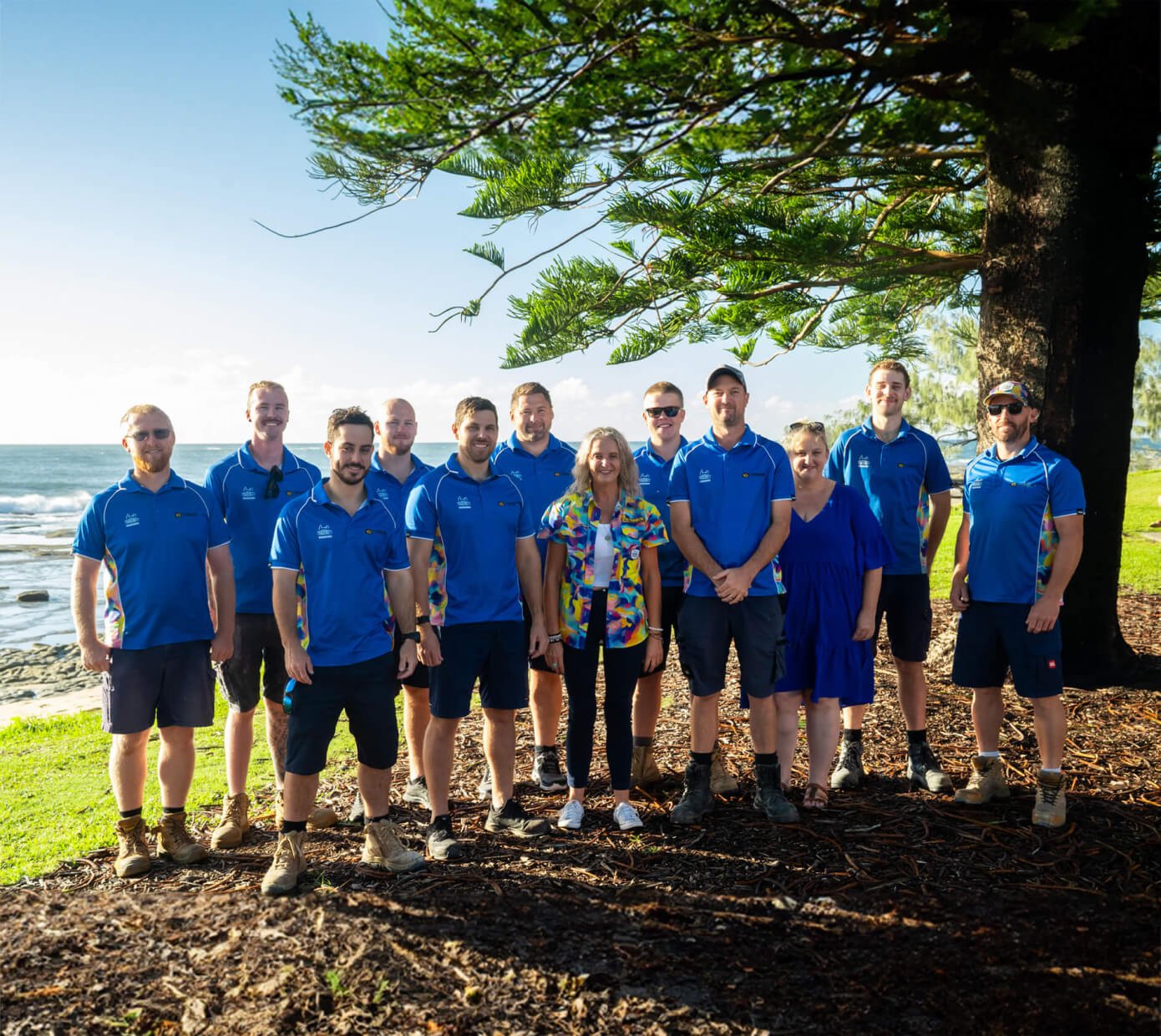 Tenmen Electrical team in blue uniforms and one in a colorful shirt stand under a tree by the seaside of the Sunshine Coast.