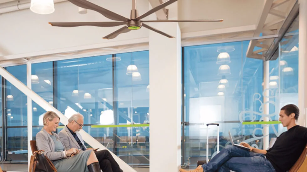 Three people sit in an airport lounge area with large windows. A ceiling fan is visible above them.