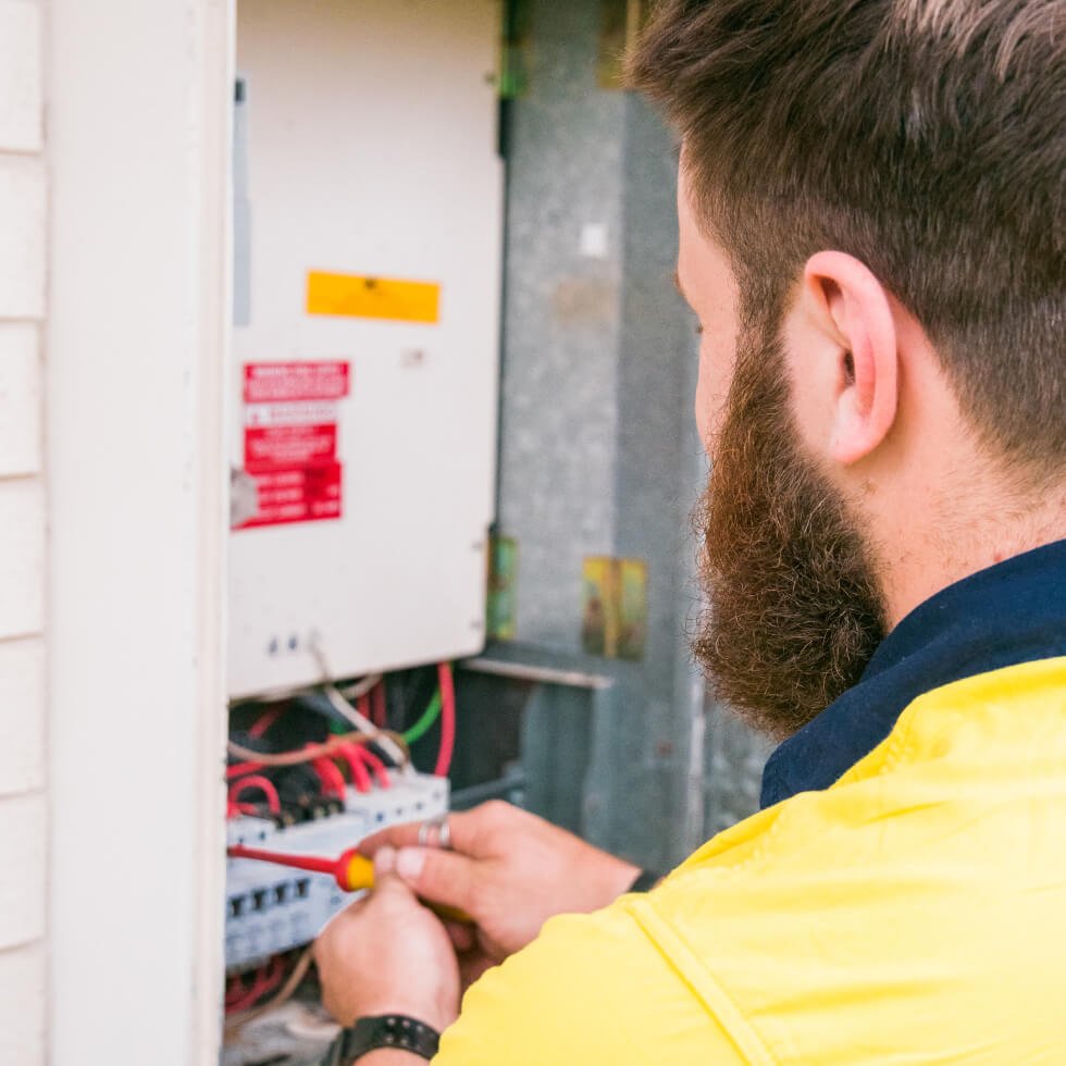 A Maroochydore electrician in a yellow shirt is working on an electrical panel.