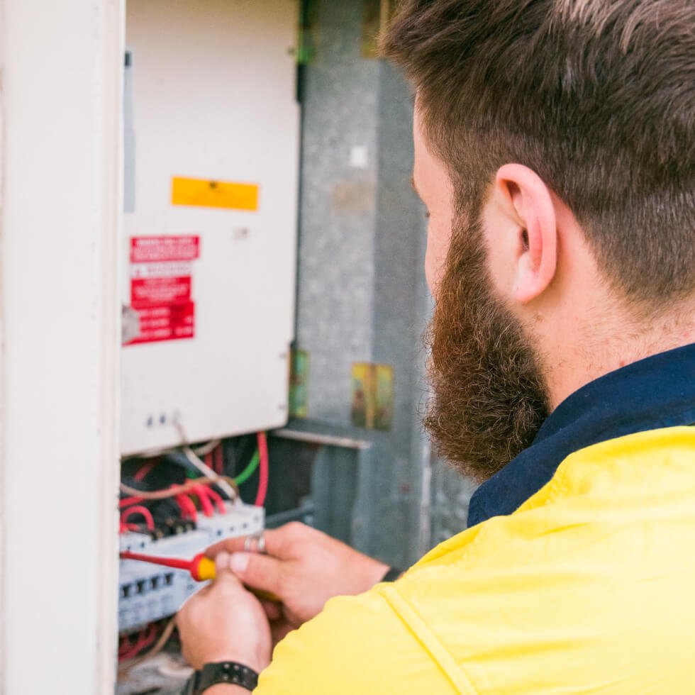 A Noosa electrician in a yellow shirt is working on an electrical panel.