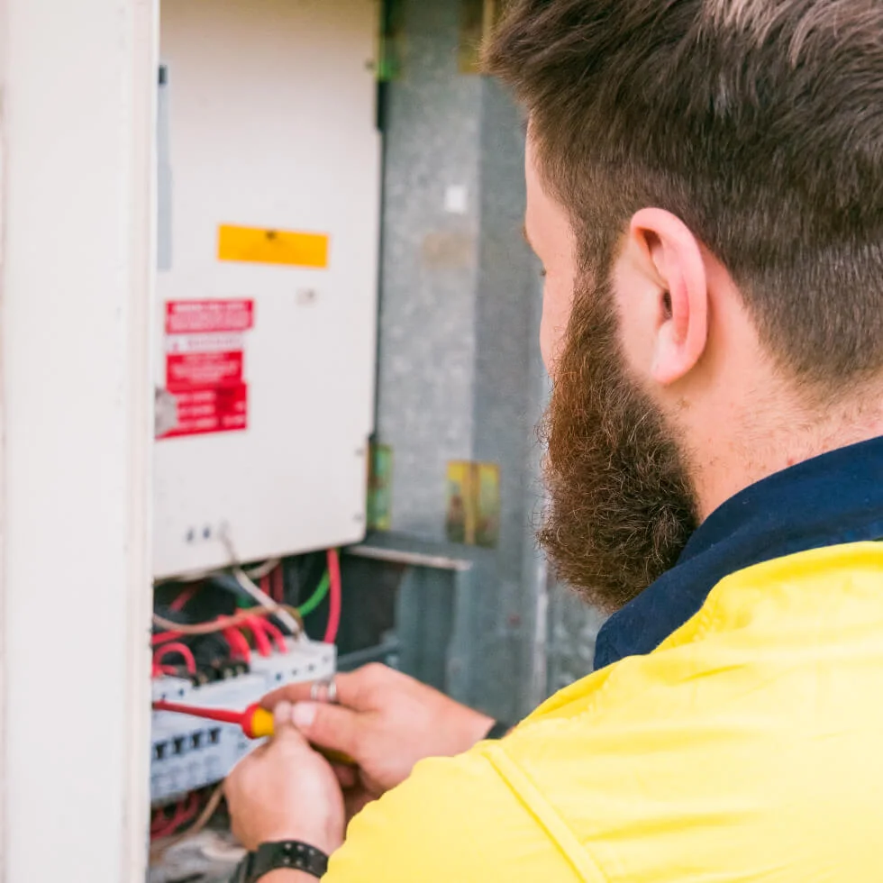 An electrician in a yellow shirt is working on an electrical panel.