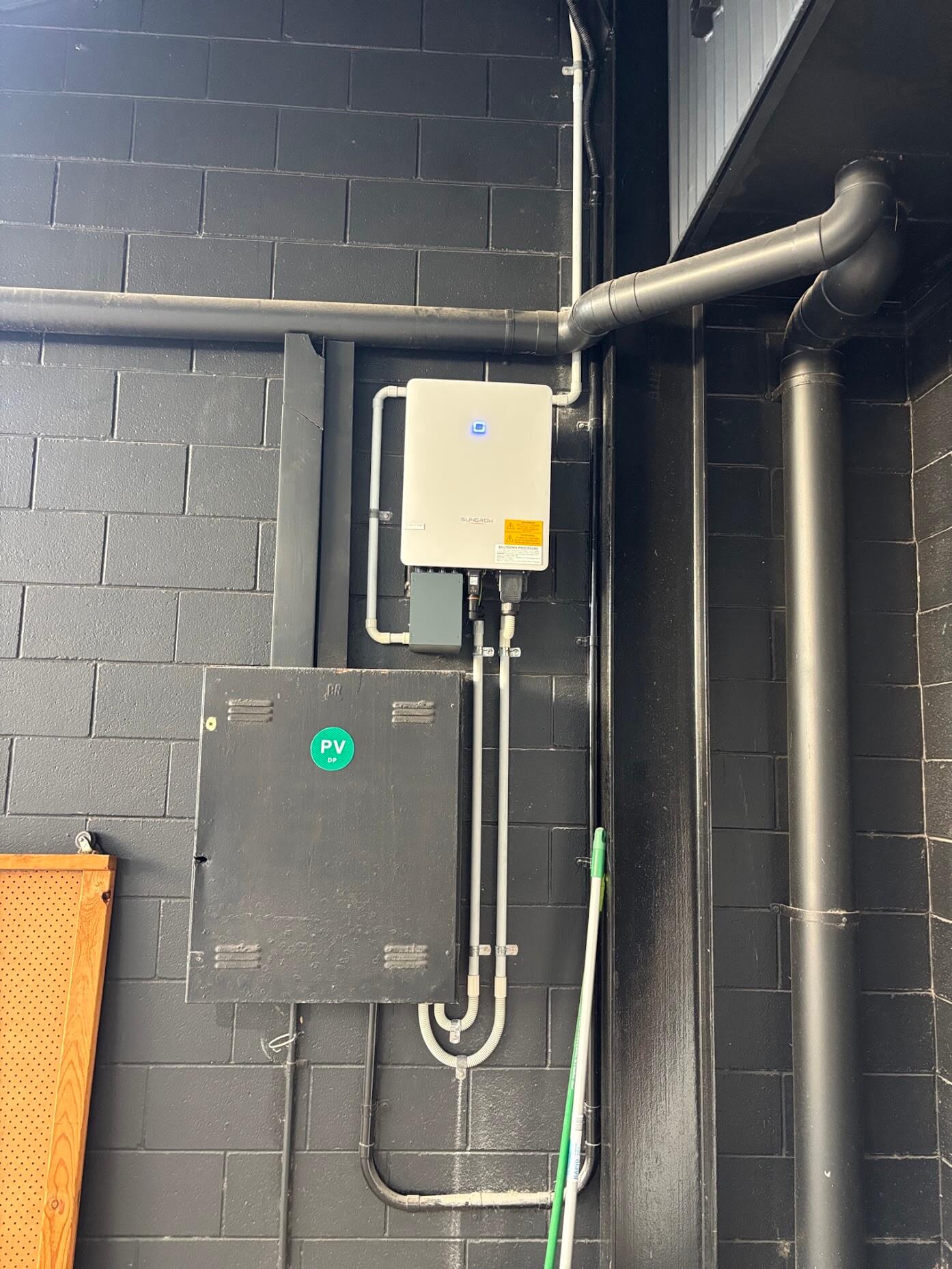 A solar inverter mounted on a black brick wall above an electrical box labeled with a green “PV” sticker.