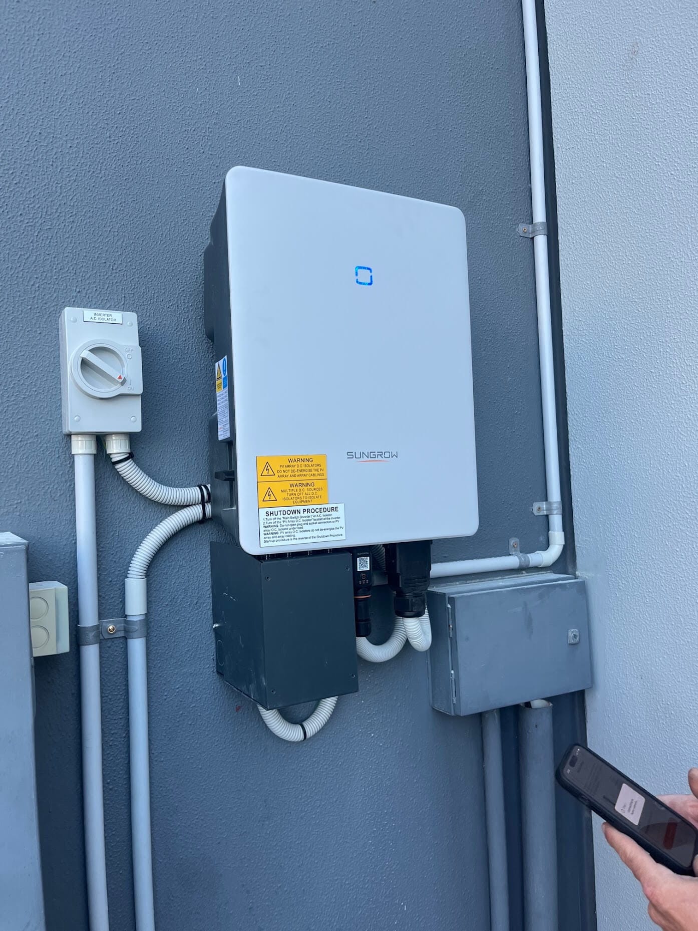 A wall-mounted Sungrow solar inverter next to an AC isolator switch, with a person holding a smartphone nearby.