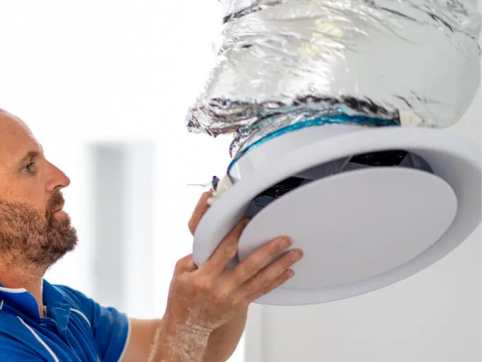 A person installs a round white ceiling vent with foil-wrapped ductwork.