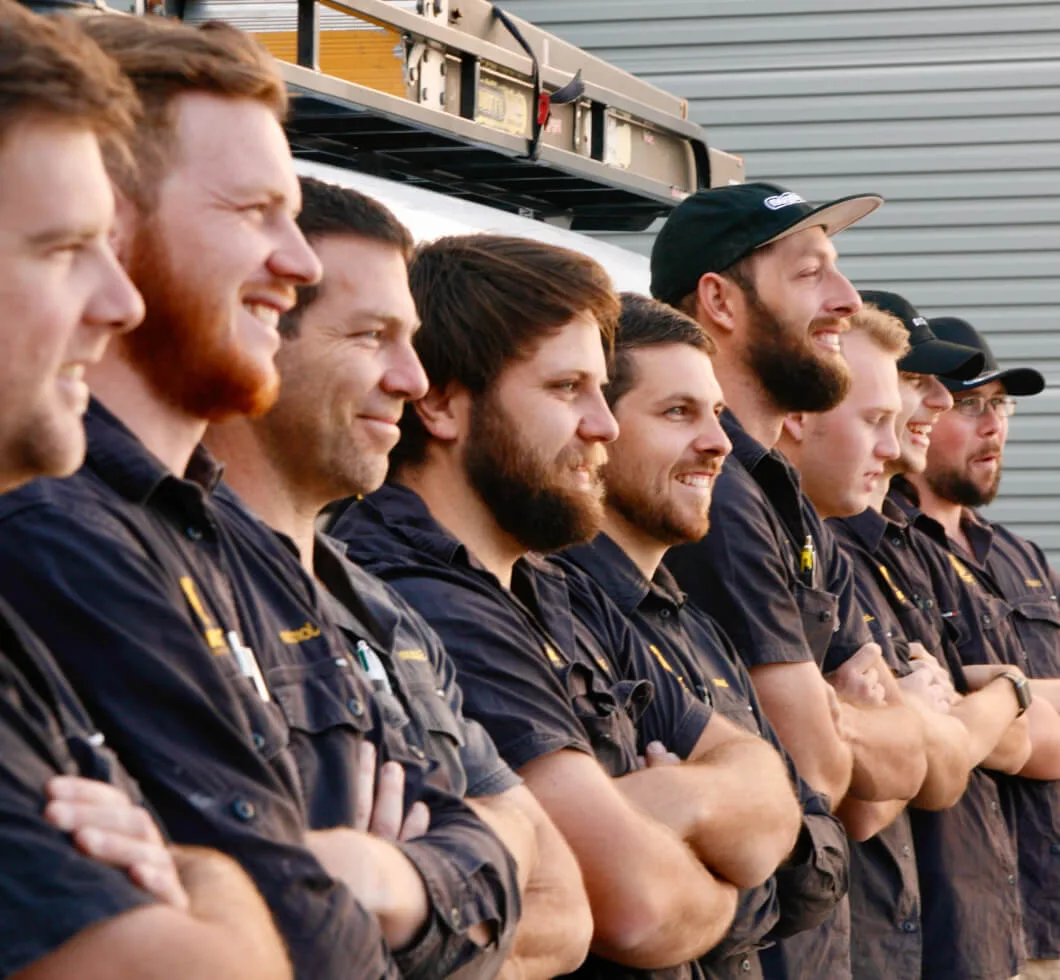 A group of men in uniform shirts stand in a row, smiling, with arms crossed. They are outdoors in front of a building with metal siding.