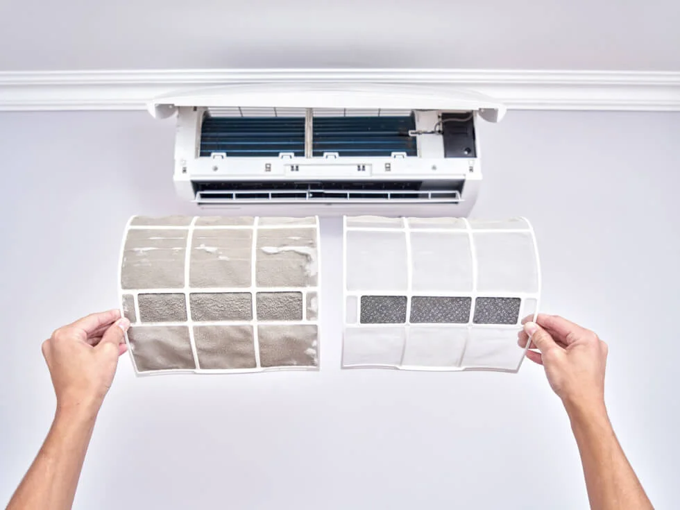 Person holding a dirty air filter next to a clean one in front of an open air conditioning unit.