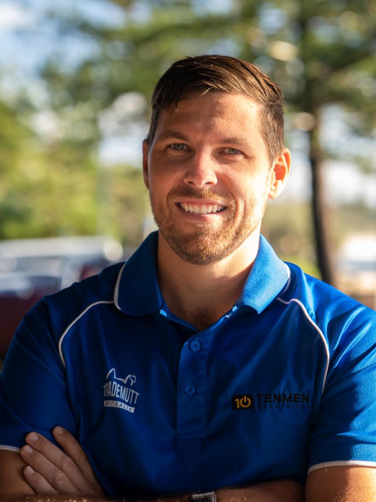 Isaac Jamieson, Tenmen Electrical director with short hair and a beard is smiling, wearing the blue uniform with logos that read "TenMen Electrical," standing outdoors with blurred greenery in the background.