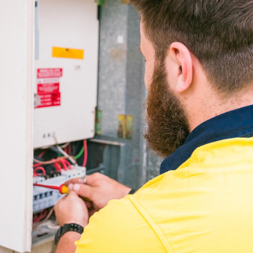 An electrician in a yellow shirt is working on an electrical panel.