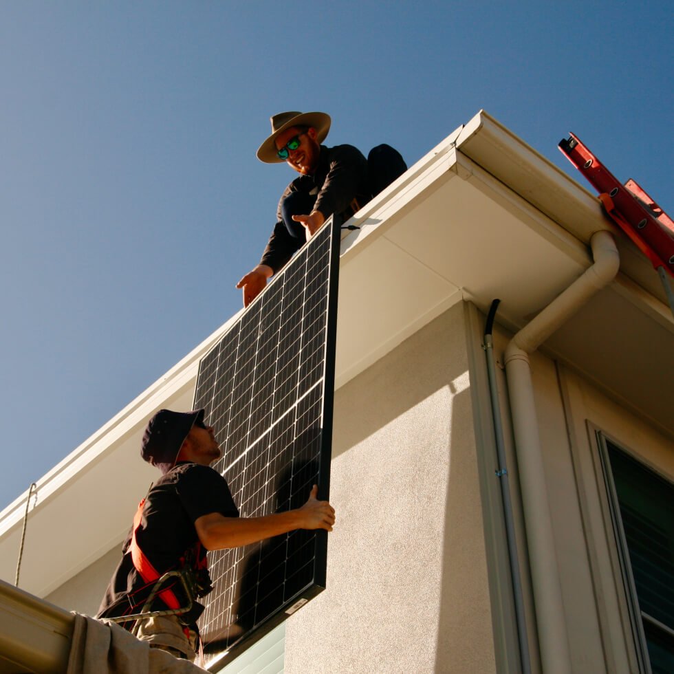 Two solar installers carrying a solar panel onto a roof of a house in Coolum