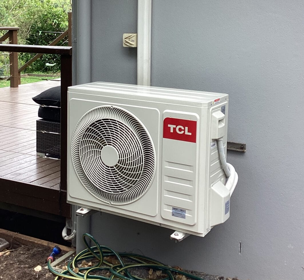 Outdoor air conditioning unit installed on a gray wall with a deck and greenery visible in the background.