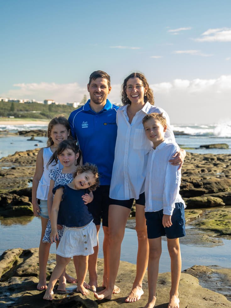 Tenmen Electrical family of five, two adults and four children, stand together on a rocky shoreline of the Sunshine Coast with the ocean in the background on a sunny day.