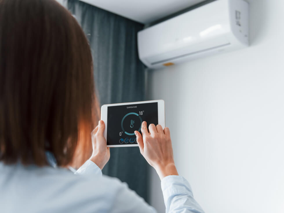 Woman adjusting a digital thermostat set to 18 degrees celsius with air conditioner unit in background.