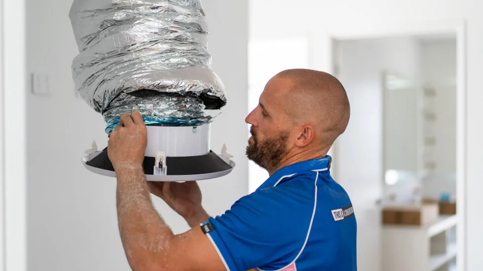 A man in a blue shirt installs a ducted air conditioning vent on a ceiling.