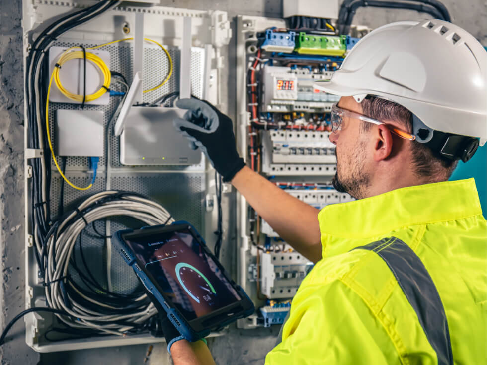 A man in a hard hat is using a tablet to check an electrical panel.
