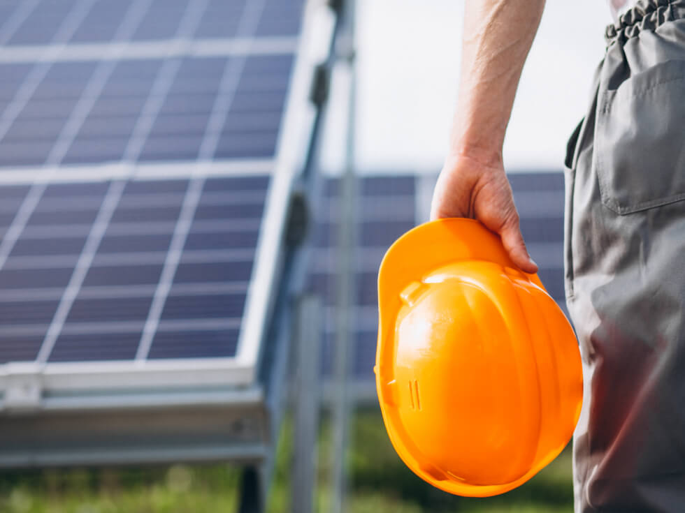 A construction worker holding a hard hat in front of solar panels.