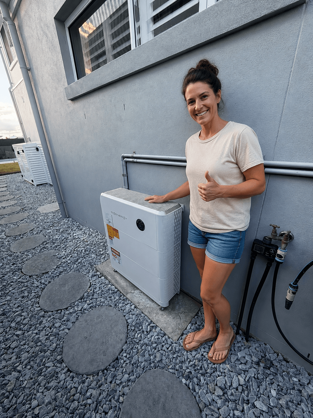 A woman stands outside by a white air conditioning unit attached to a gray building, giving a thumbs up and smiling.