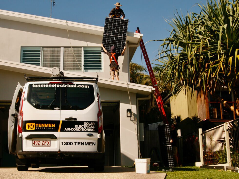 The Tenmen van parked in front of a house and two solar installers carrying a solar panel on the roof.